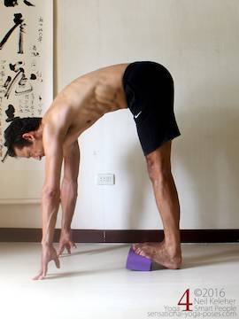 Standing calf stretch while bending forwards, fronts of feet elevated with yoga block. Hips forwards to reduce stretch.